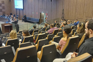 A imagem mostra o grande auditório onde ocorrem as reuniões do Órgão Especial do TJMG. Há um grupo de pessoas (desembargadores) reunidas à frente, em uma grande mesa com um telão ao fundo. Nas cadeiras enfileiradas, há diversas pessoas acompanhando a reunião, entre elas está o diretor de Assuntos Jurídicos do SINJUS-MG, Felipe Rodrigues.