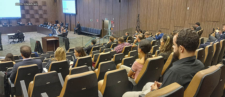 A imagem mostra o grande auditório onde ocorrem as reuniões do Órgão Especial do TJMG. Há um grupo de pessoas (desembargadores) reunidas à frente, em uma grande mesa com um telão ao fundo. Nas cadeiras enfileiradas, há diversas pessoas acompanhando a reunião, entre elas está o diretor de Assuntos Jurídicos do SINJUS-MG, Felipe Rodrigues.