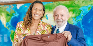Fotografia tirada em uma sala de reuniões no Palácio do Planalto, na foto se vê a ministra Anielle Franco, com fisionomia alegre ao lado do presidente Lula com o mesmo semblante de alegria, ambos estão segurandp uma camiseta na cor marrom com a seguinte frase estampada "Brasil pela igualdade racial".