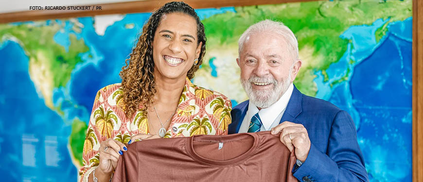 Fotografia tirada em uma sala de reuniões no Palácio do Planalto, na foto se vê a ministra Anielle Franco, com fisionomia alegre ao lado do presidente Lula com o mesmo semblante de alegria, ambos estão segurandp uma camiseta na cor marrom com a seguinte frase estampada "Brasil pela igualdade racial".