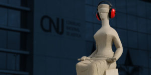 Escultura da Justiça em frente ao prédio do Conselho Nacional de Justiça (CNJ), com o fundo desfocado mostrando o logotipo da instituição. A escultura, representando uma figura feminina de olhos vendados e sentada com uma espada no colo, aparece editada com fones de ouvido vermelhos de proteção acústica, sugerindo uma crítica visual à surdez institucional. Foto original por Romulo Serpa/Agência CNJ.