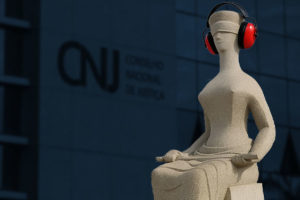 Escultura da Justiça em frente ao prédio do Conselho Nacional de Justiça (CNJ), com o fundo desfocado mostrando o logotipo da instituição. A escultura, representando uma figura feminina de olhos vendados e sentada com uma espada no colo, aparece editada com fones de ouvido vermelhos de proteção acústica, sugerindo uma crítica visual à surdez institucional. Foto original por Romulo Serpa/Agência CNJ.