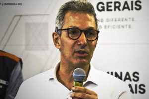 Imagem Acessível: Homem branco e cabelos grisalhos fala usando microfone, é Romeu Zema, governador de Minas Gerais. A imagem está com tratamento que dá um ar sombrio.