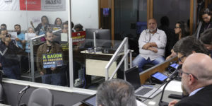 Servidores acompanham a votação do PL do Auxílio-Saúde na ALMG, vistos por uma divisória de vidro. Alguns seguram cartazes com frases como “A saúde do servidor também importa” e “Auxílio-saúde digno já”. Entre os servidores, em primeiro plano estão os diretores do SINJUS, Felipe Rodrigues (diretoria de Assuntos Jurídicos) e Alexandre Gomes (diretoria de Imprensa e Comunicação).