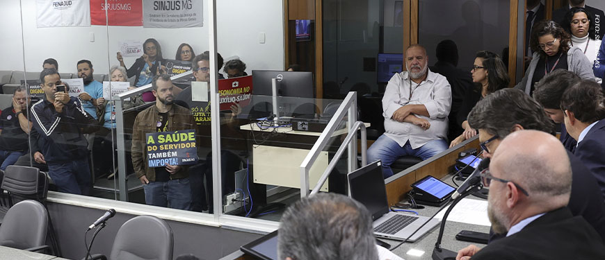 Servidores acompanham a votação do PL do Auxílio-Saúde na ALMG, vistos por uma divisória de vidro. Alguns seguram cartazes com frases como “A saúde do servidor também importa” e “Auxílio-saúde digno já”. Entre os servidores, em primeiro plano estão os diretores do SINJUS, Felipe Rodrigues (diretoria de Assuntos Jurídicos) e Alexandre Gomes (diretoria de Imprensa e Comunicação).