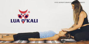 Imagem Acessível: A imagem mostra uma sessão de Reiki, na cena, há duas mulheres: uma terapeuta e uma paciente. A terapeuta, à esquerda, está sentada em uma almofada, vestindo uma blusa preta sem mangas e calça preta. Ela tem cabelos longos e está inclinada para a frente, com as mãos posicionadas sob a paciente. A paciente está deitada de costas em um tapete,com os braços relaxados ao longo do corpo. Ela está vestindo uma camiseta azul e calças justas pretas. O ambiente é simples e acolhedor, com piso de madeira e paredes brancas.