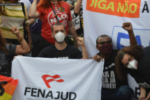 #ImagemAcessível: Foto de uma manifestação contra a PEC 32. No centro da foto está o coordenador-geral do SINJUS, Alexandre Pires: homem branco de olhos castanhos, cabelos curtos e castanhos, usando uma máscara de proteção branca, camisa preta. Com uma das mãos ele segura uma faixa branca com a logomarca da Fenajud e a outra está levantada com o punho cerrado. Ao lado esquerdo dele está uma mulher de cabelos longos, cacheados de cor castanho claro, usando máscara de proteção preta e blusa preta com os dizeres: lute como uma professora. Ela também segura uma faixa colorida e está com o punho cerrado. Do lado direito está um homem negro de cabelos pretos e curtos, olhos castanhos, usando blusa preta, óculos escuros e máscara de proteção preta. Ele segura a mesma faixa que Alexandre e também está com o punho cerrado. Ao lado dele está uma mulher branca com cabelos longos de cor castanho claro e olhos castanhos. Ela usa uma máscara de proteção branca e uma blusa preta. No fundo, observa-se faixas e uma multidão com blusas e faixas com dizeres contra a PEC 32.