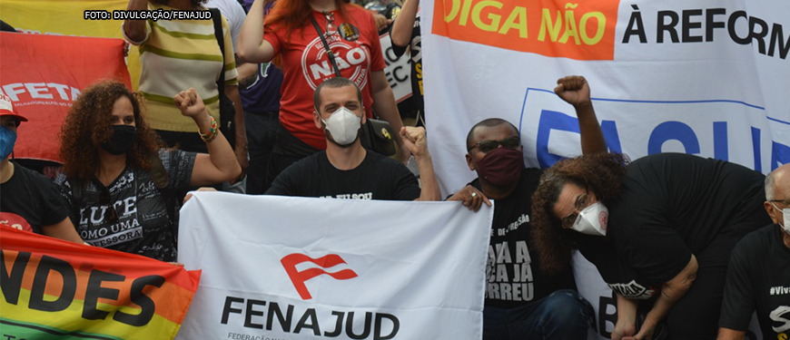 #ImagemAcessível: Foto de uma manifestação contra a PEC 32. No centro da foto está o coordenador-geral do SINJUS, Alexandre Pires: homem branco de olhos castanhos, cabelos curtos e castanhos, usando uma máscara de proteção branca, camisa preta. Com uma das mãos ele segura uma faixa branca com a logomarca da Fenajud e a outra está levantada com o punho cerrado. Ao lado esquerdo dele está uma mulher de cabelos longos, cacheados de cor castanho claro, usando máscara de proteção preta e blusa preta com os dizeres: lute como uma professora. Ela também segura uma faixa colorida e está com o punho cerrado. Do lado direito está um homem negro de cabelos pretos e curtos, olhos castanhos, usando blusa preta, óculos escuros e máscara de proteção preta. Ele segura a mesma faixa que Alexandre e também está com o punho cerrado. Ao lado dele está uma mulher branca com cabelos longos de cor castanho claro e olhos castanhos. Ela usa uma máscara de proteção branca e uma blusa preta. No fundo, observa-se faixas e uma multidão com blusas e faixas com dizeres contra a PEC 32.