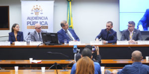 Registro de uma audiência pública na Câmara dos Deputados. Três homens estão sentados à mesa principal, com microfones e crachás de identificação. Ao fundo, há uma faixa com o texto “Audiência Pública — 20 anos do CNJ. Por um Judiciário mais democrático e representativo.” e uma bandeira do Brasil. À frente da mesa, o público acompanha a sessão. O homem ao centro, de terno azul, é o deputado que preside a audiência; à direita, o coordenador-geral do SINJUS-MG, Alexandre Pires, fala ao microfone, gesticulando com as mãos.