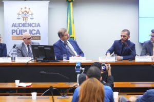 Registro de uma audiência pública na Câmara dos Deputados. Três homens estão sentados à mesa principal, com microfones e crachás de identificação. Ao fundo, há uma faixa com o texto “Audiência Pública — 20 anos do CNJ. Por um Judiciário mais democrático e representativo.” e uma bandeira do Brasil. À frente da mesa, o público acompanha a sessão. O homem ao centro, de terno azul, é o deputado que preside a audiência; à direita, o coordenador-geral do SINJUS-MG, Alexandre Pires, fala ao microfone, gesticulando com as mãos.