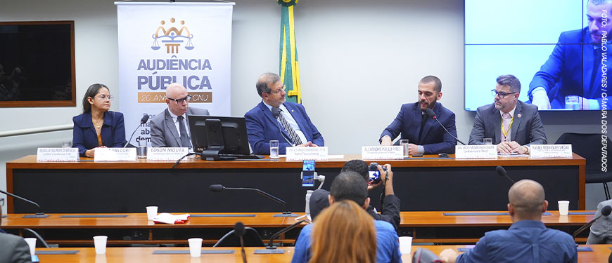 Registro de uma audiência pública na Câmara dos Deputados. Três homens estão sentados à mesa principal, com microfones e crachás de identificação. Ao fundo, há uma faixa com o texto “Audiência Pública — 20 anos do CNJ. Por um Judiciário mais democrático e representativo.” e uma bandeira do Brasil. À frente da mesa, o público acompanha a sessão. O homem ao centro, de terno azul, é o deputado que preside a audiência; à direita, o coordenador-geral do SINJUS-MG, Alexandre Pires, fala ao microfone, gesticulando com as mãos.