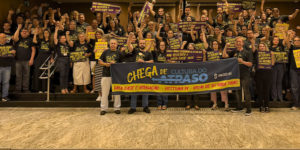 Registro de uma mobilização de dezenas de servidores no hall do Órgão Especial do TJMG. Os participantes estão reunidos na escadaria, com os punhos erguidos, vestindo camisetas pretas e segurando cartazes de protesto. À frente, está o coordenador-geral do SINJUS-MG, Felipe Rodrigues junto dos suplentes Robert França e Augusto Chagas exibem uma faixa com a mensagem “Chega de Cultura do Atraso”, reivindicando Data-Base, Promoção Vertical e opção de jornada de 8 horas para todos.