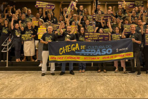 Registro de uma mobilização de dezenas de servidores no hall do Órgão Especial do TJMG. Os participantes estão reunidos na escadaria, com os punhos erguidos, vestindo camisetas pretas e segurando cartazes de protesto. À frente, está o coordenador-geral do SINJUS-MG, Felipe Rodrigues junto dos suplentes Robert França e Augusto Chagas exibem uma faixa com a mensagem “Chega de Cultura do Atraso”, reivindicando Data-Base, Promoção Vertical e opção de jornada de 8 horas para todos.