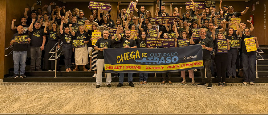 Registro de uma mobilização de dezenas de servidores no hall do Órgão Especial do TJMG. Os participantes estão reunidos na escadaria, com os punhos erguidos, vestindo camisetas pretas e segurando cartazes de protesto. À frente, está o coordenador-geral do SINJUS-MG, Felipe Rodrigues junto dos suplentes Robert França e Augusto Chagas exibem uma faixa com a mensagem “Chega de Cultura do Atraso”, reivindicando Data-Base, Promoção Vertical e opção de jornada de 8 horas para todos.