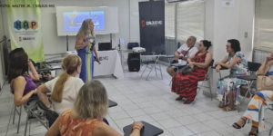 Imagem Acessível: A imagem retrata um grupo de pessoas reunidas, participantes do núcleo NAP em uma sala, sentadas em cadeiras e de frente para a palestrante, uma mulher branca de cabelos loiros, que está parada em frente a um quadro branco. O público é engajado e atento, e ao fundo se vê o banner com a logo do NAP.