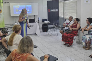 Imagem Acessível: A imagem retrata um grupo de pessoas reunidas, participantes do núcleo NAP em uma sala, sentadas em cadeiras e de frente para a palestrante, uma mulher branca de cabelos loiros, que está parada em frente a um quadro branco. O público é engajado e atento, e ao fundo se vê o banner com a logo do NAP.