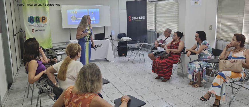 Imagem Acessível: A imagem retrata um grupo de pessoas reunidas, participantes do núcleo NAP em uma sala, sentadas em cadeiras e de frente para a palestrante, uma mulher branca de cabelos loiros, que está parada em frente a um quadro branco. O público é engajado e atento, e ao fundo se vê o banner com a logo do NAP.