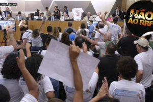 Pessoas em pé levantando cartazes com palavras de ordem contra o RRF, elas estão com punhos para o alto em gesto de luta e alguns apontam para deputados da mesa em auditório da ALMG, onde ocorreu a Audiência Pública sobre o RRF, Regime de Recuperação Fiscal. Conteúdo textual: PREJUÍZOS - DEPUTADOS E SINDICALISTAS DESMENTEM GOVERNO EM AUDIÊNCIA PÚBLICA SOBRE IMPACTOS DO RRF.