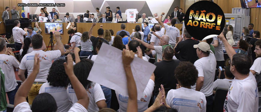 Pessoas em pé levantando cartazes com palavras de ordem contra o RRF, elas estão com punhos para o alto em gesto de luta e alguns apontam para deputados da mesa em auditório da ALMG, onde ocorreu a Audiência Pública sobre o RRF, Regime de Recuperação Fiscal. Conteúdo textual: PREJUÍZOS - DEPUTADOS E SINDICALISTAS DESMENTEM GOVERNO EM AUDIÊNCIA PÚBLICA SOBRE IMPACTOS DO RRF.