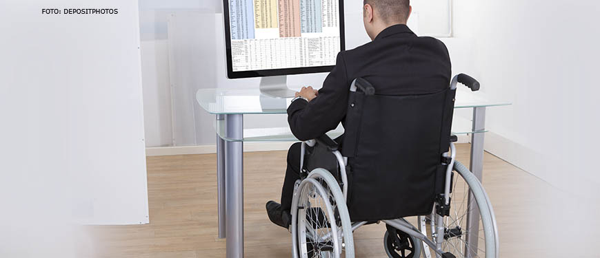 Pessoa com deficiência em uma cadeira de rodas trabalha diante de um computador em um escritório, ela está de costas e é possível ver uma grande planilha na tela de seu computador.