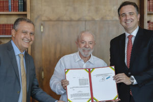 A imagem mostra três autoridades sorridentes em um ambiente formal, com estantes de livros ao fundo. O centro da cena é ocupado por um documento oficial, com destaque para a assinatura. O texto sobreposto informa: "Propag é regulamentado e Minas deve decidir sobre estatais até outubro", com ênfase na parte “Propag é regulamentado” em vermelho, destacando a importância da medida.