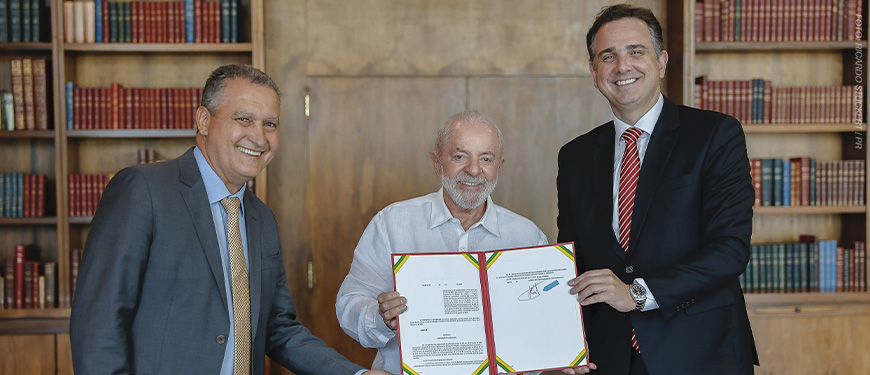 A imagem mostra três autoridades sorridentes em um ambiente formal, com estantes de livros ao fundo. O centro da cena é ocupado por um documento oficial, com destaque para a assinatura. O texto sobreposto informa: "Propag é regulamentado e Minas deve decidir sobre estatais até outubro", com ênfase na parte “Propag é regulamentado” em vermelho, destacando a importância da medida.