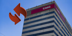 Imagem Acessível: Imagem em formato vertical de um prédio institucional alto, visto de baixo para cima, contra um céu azul limpo. No topo da edificação, há uma faixa vermelha com a sigla “TJMG” em destaque. A fachada é composta por faixas horizontais claras intercaladas com janelas escuras espelhadas. Na base da imagem, aparecem árvores verdes e alguns fios elétricos, indicando que a foto foi feita em área urbana. À direita da imagem, há um elemento gráfico em vermelho com duas setas curvas, sugerindo movimento ou troca. Conteúdo textual: LOTAÇÃO - TJMG convoca servidores classificados no Edital de Remoção nº 01/2025.