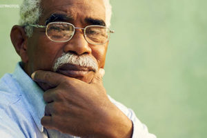 Homem negro com cabelos brancos e curtos está em pose reflexiva, com a mão no queixo e semblante sério. Ele usa óculos e uma camisa jeans.