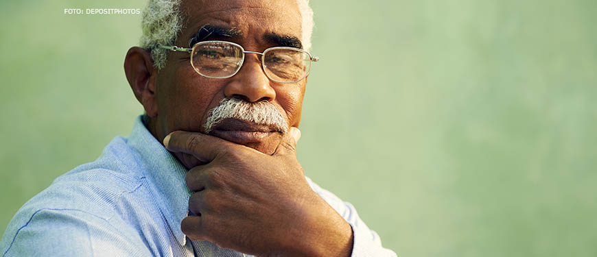 Homem negro com cabelos brancos e curtos está em pose reflexiva, com a mão no queixo e semblante sério. Ele usa óculos e uma camisa jeans.