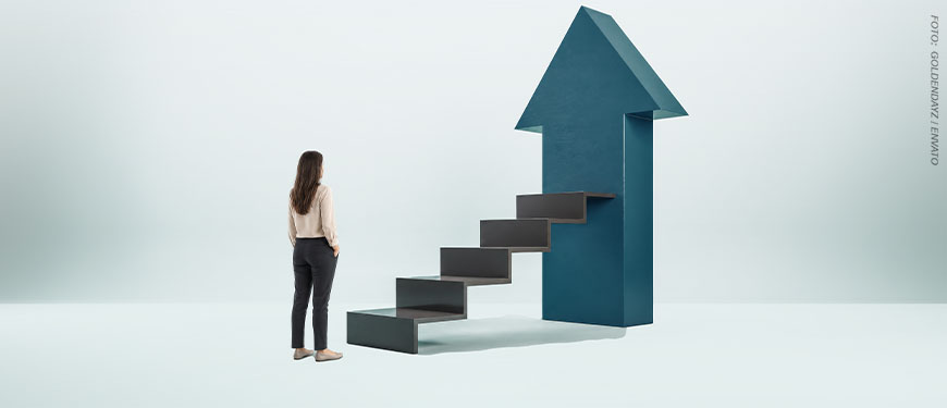 Imagem Acessível: Imagem com visual minimalista em tons de cinza e azul, nela há uma mulher vista de costas, posicionada à esquerda. Ela tem cabelos castanhos longos, veste camisa clara de mangas compridas, calça social escura e sapatos discretos. A mulher observa uma escada escura que sobe em direção a uma grande seta azul apontada para cima. A escada está integrada à estrutura da seta, simbolizando crescimento, progresso e ascensão na carreira. A seta ocupa grande parte do lado direito da composição e se destaca sobre um fundo claro e neutro. Contéudo textual: Carreira - TJMG divulga resultado preliminar da PV 2024.