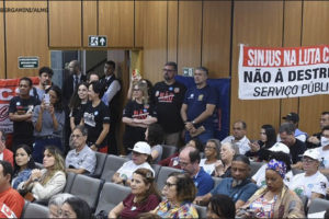 Foto de auditório na Assembleia Legislativa de Minas Gerais, com a Comissão da Administração Pública, onde há diversas pessoas sentadas e algumas em pé. Em destaque, está a faixa com o seguinte texto: "Sinjus na luta contra a RRF – Não à destruição do serviço público!", e alguns dirigentes do Sindicato estão ao lado dela.