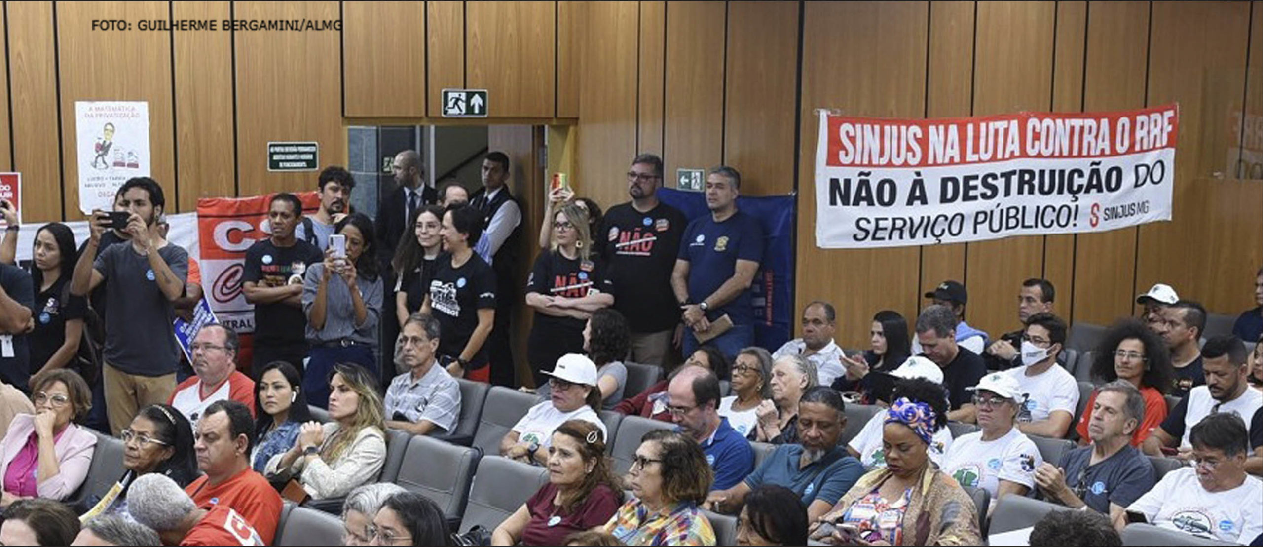 Foto de auditório na Assembleia Legislativa de Minas Gerais, com a Comissão da Administração Pública, onde há diversas pessoas sentadas e algumas em pé. Em destaque, está a faixa com o seguinte texto: "Sinjus na luta contra a RRF – Não à destruição do serviço público!", e alguns dirigentes do Sindicato estão ao lado dela.