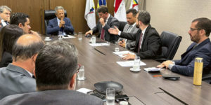 Fotografia de reunião entre representantes sindicais e o presidente do TJMG, o desembargador Vicente de Oliveira Silva, também estão presentes integrantes da alta administração do TJMG. Os participantes estão sentados ao redor de uma mesa, ao fundo há um suporte com bandeiras. O coordenador-geral do SINJUS-MG, Felipe Rodrigues está vestido com um terno escuro e gravata listrada. Ele fala e gesticula enquanto o presidente do TJMG e os demais acompanham. Os outros participantes são os dirigentes sindicais do Serjusmig e Sindojus/MG.