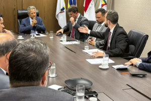 Fotografia de reunião entre representantes sindicais e o presidente do TJMG, o desembargador Vicente de Oliveira Silva, também estão presentes integrantes da alta administração do TJMG. Os participantes estão sentados ao redor de uma mesa, ao fundo há um suporte com bandeiras. O coordenador-geral do SINJUS-MG, Felipe Rodrigues está vestido com um terno escuro e gravata listrada. Ele fala e gesticula enquanto o presidente do TJMG e os demais acompanham. Os outros participantes são os dirigentes sindicais do Serjusmig e Sindojus/MG.