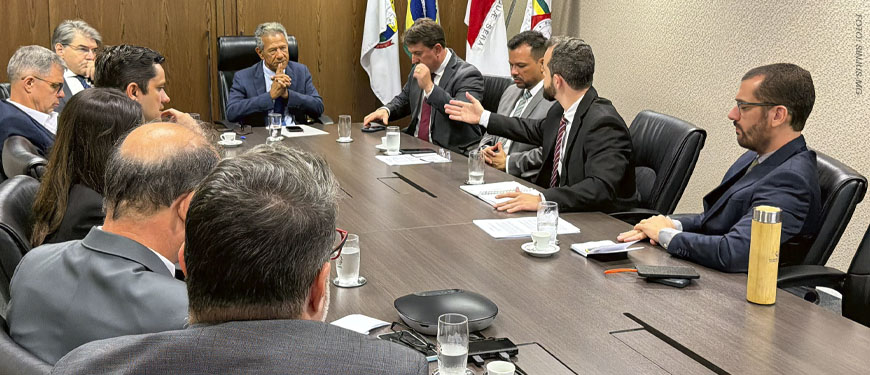 Fotografia de reunião entre representantes sindicais e o presidente do TJMG, o desembargador Vicente de Oliveira Silva, também estão presentes integrantes da alta administração do TJMG. Os participantes estão sentados ao redor de uma mesa, ao fundo há um suporte com bandeiras. O coordenador-geral do SINJUS-MG, Felipe Rodrigues está vestido com um terno escuro e gravata listrada. Ele fala e gesticula enquanto o presidente do TJMG e os demais acompanham. Os outros participantes são os dirigentes sindicais do Serjusmig e Sindojus/MG.