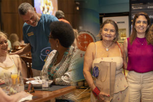 A imagem reúne dois registros da confraternização do NAP e do NPD do SINJUS, em um ambiente descontraído, com pessoas sorrindo e interagindo em mesas. Na parte superior, o diretor Alex Aguiar cumprimenta duas filiadas com um abraço amistoso. Na parte inferior, a diretora Janaína Torres aparece ao lado de duas filiadas, todas sorridentes e segurando lembranças do evento.