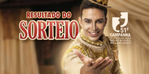 Imagem Acessível: A imagem mostra um ator vestido com um figurino teatral dourado e uma coroa ornamentada, estendendo a mão em direção ao público, em um cenário com cortinas de palco ao fundo. O texto em destaque, centralizado na parte superior, diz: “Resultado do Sorteio”, em letras grandes e vermelhas. Logo abaixo, está o logotipo da 51ª Campanha de Popularização do Teatro & Dança de Belo Horizonte, seguido do logotipo do SINJUS-MG, que promoveu o sorteio dos vouchers culturais.