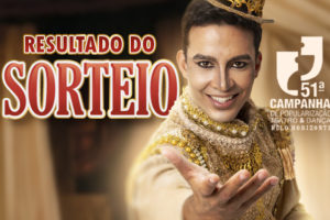 Imagem Acessível: A imagem mostra um ator vestido com um figurino teatral dourado e uma coroa ornamentada, estendendo a mão em direção ao público, em um cenário com cortinas de palco ao fundo. O texto em destaque, centralizado na parte superior, diz: “Resultado do Sorteio”, em letras grandes e vermelhas. Logo abaixo, está o logotipo da 51ª Campanha de Popularização do Teatro & Dança de Belo Horizonte, seguido do logotipo do SINJUS-MG, que promoveu o sorteio dos vouchers culturais.
