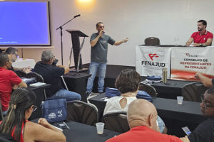 A imagem retrata um auditório com servidoras e servidores sentados em fileiras, acompanhando um encontro sindical. Ao centro, o coordenador-geral do SINJUS e coordenador de Assuntos Jurídicos da Fenajud, Alexandre Pires, fala ao microfone e gesticula para o público. Há uma tela de projeção à esquerda e elementos gráficos sobrepostos destacando a temática da articulação nacional, além da marca do SINJUS-MG na parte inferior.