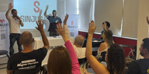 Imagem acessível: A imagem mostra o ambiente interno na Sede do SINJUS-MG, com várias pessoas sentadas em cadeiras, com as mãos levantadas em sinal de votação; à frente, dois dirigentes do sindicato estão próximos a uma mesa e a um telão com a identidade do SINJUS-MG, conduzindo a reunião. No topo da imagem, aparece a versal “Transparência” e, sobre a imagem, o texto em destaque diz: “AGO aprova contas de 2025 e previsão orçamentária para 2026”.