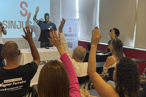Imagem acessível: A imagem mostra o ambiente interno na Sede do SINJUS-MG, com várias pessoas sentadas em cadeiras, com as mãos levantadas em sinal de votação; à frente, dois dirigentes do sindicato estão próximos a uma mesa e a um telão com a identidade do SINJUS-MG, conduzindo a reunião. No topo da imagem, aparece a versal “Transparência” e, sobre a imagem, o texto em destaque diz: “AGO aprova contas de 2025 e previsão orçamentária para 2026”.