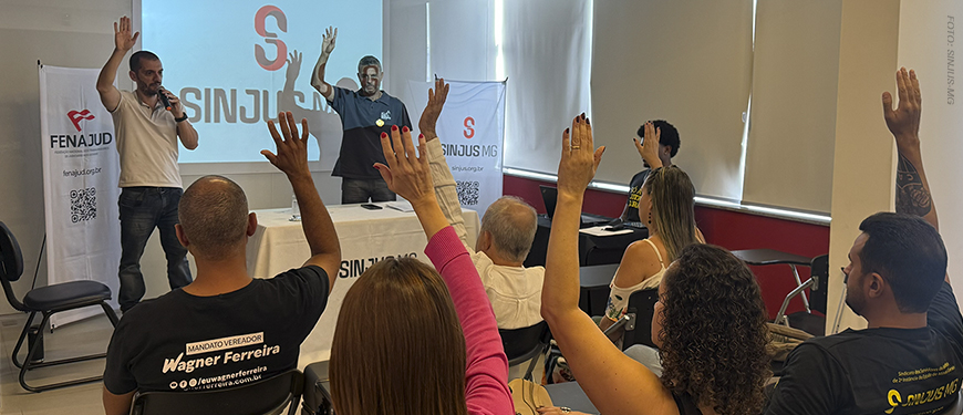 Imagem acessível: A imagem mostra o ambiente interno na Sede do SINJUS-MG, com várias pessoas sentadas em cadeiras, com as mãos levantadas em sinal de votação; à frente, dois dirigentes do sindicato estão próximos a uma mesa e a um telão com a identidade do SINJUS-MG, conduzindo a reunião. No topo da imagem, aparece a versal “Transparência” e, sobre a imagem, o texto em destaque diz: “AGO aprova contas de 2025 e previsão orçamentária para 2026”.