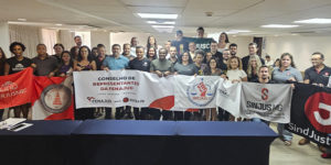 A imagem mostra um grupo numeroso de representantes sindicais posando para foto coletiva durante o Conselho de Representantes da Fenajud. Várias pessoas seguram faixas e bandeiras de seus sindicatos, incluindo o SINJUS-MG.