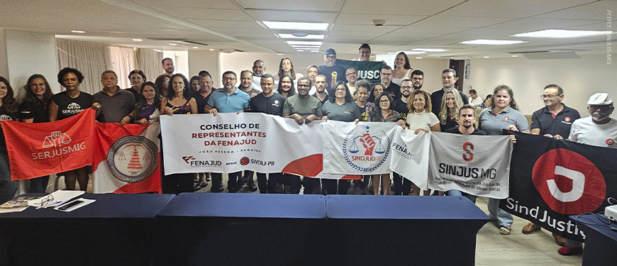 A imagem mostra um grupo numeroso de representantes sindicais posando para foto coletiva durante o Conselho de Representantes da Fenajud. Várias pessoas seguram faixas e bandeiras de seus sindicatos, incluindo o SINJUS-MG.