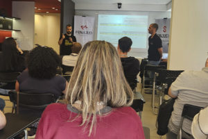 Servidores sentados em uma sala assistem a uma apresentação durante reunião ampliada do SINJUS. O diretor Felipe Rodrigues fala ao microfone em pé, ao lado de uma tela com projeção. A pauta é a mobilização por mais vagas na Promoção Vertical de 2023 (PV 2023).