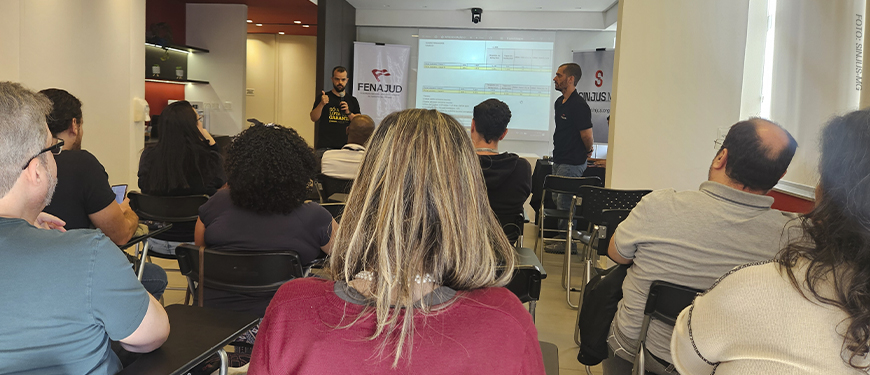 Servidores sentados em uma sala assistem a uma apresentação durante reunião ampliada do SINJUS. O diretor Felipe Rodrigues fala ao microfone em pé, ao lado de uma tela com projeção. A pauta é a mobilização por mais vagas na Promoção Vertical de 2023 (PV 2023).
