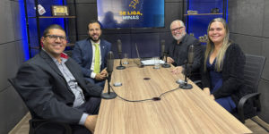 Fotografia registrada em um estúdio de podcast. Quatro pessoas estão sentadas ao redor de uma mesa retangular de madeira, cada uma com um microfone à sua frente. Da esquerda para a direita, um homem de óculos veste blazer escuro e camisa cinza ( diretor do Sindicato dos Servidores do Ministério Público do Estado de Minas Gerais (Sindsemp-MG) Enildo Gomes Meira Filho, ; ao lado, um homem de terno azul, camisa branca e gravata verde sorri para a câmera (Associação das Defensoras e Defensores Públicos de Minas Gerais (ADEP-MG), Rômulo Veloso de Carvalho); em seguida, um homem de barba branca, óculos e camisa preta também sorri (o jornalista Orion Teixeira, mediador do programa); à direita, uma mulher de cabelos longos loiros veste blusa azul e casaco preto (a diretora suplente do SINJUS-MG, Flávia Maria da Silveira). Ao fundo, um monitor exibe a logotipo do programa “Se Liga, Minas”, composta pelo contorno do mapa de Minas Gerais e um sino estilizado. O estúdio tem paredes com tratamento acústico, iluminação em tons de azul e estantes laterais com objetos decorativos, como um rádio antigo, câmeras, livros e itens de comunicação. Sobre a mesa há um notebook, copos com água e cabos conectando os microfones.