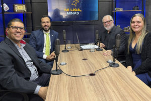 Fotografia registrada em um estúdio de podcast. Quatro pessoas estão sentadas ao redor de uma mesa retangular de madeira, cada uma com um microfone à sua frente. Da esquerda para a direita, um homem de óculos veste blazer escuro e camisa cinza ( diretor do Sindicato dos Servidores do Ministério Público do Estado de Minas Gerais (Sindsemp-MG) Enildo Gomes Meira Filho, ; ao lado, um homem de terno azul, camisa branca e gravata verde sorri para a câmera (Associação das Defensoras e Defensores Públicos de Minas Gerais (ADEP-MG), Rômulo Veloso de Carvalho); em seguida, um homem de barba branca, óculos e camisa preta também sorri (o jornalista Orion Teixeira, mediador do programa); à direita, uma mulher de cabelos longos loiros veste blusa azul e casaco preto (a diretora suplente do SINJUS-MG, Flávia Maria da Silveira). Ao fundo, um monitor exibe a logotipo do programa “Se Liga, Minas”, composta pelo contorno do mapa de Minas Gerais e um sino estilizado. O estúdio tem paredes com tratamento acústico, iluminação em tons de azul e estantes laterais com objetos decorativos, como um rádio antigo, câmeras, livros e itens de comunicação. Sobre a mesa há um notebook, copos com água e cabos conectando os microfones.