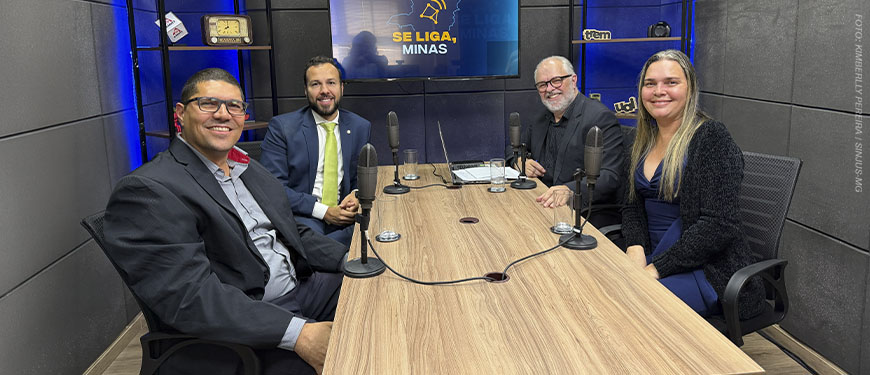 Fotografia registrada em um estúdio de podcast. Quatro pessoas estão sentadas ao redor de uma mesa retangular de madeira, cada uma com um microfone à sua frente. Da esquerda para a direita, um homem de óculos veste blazer escuro e camisa cinza ( diretor do Sindicato dos Servidores do Ministério Público do Estado de Minas Gerais (Sindsemp-MG) Enildo Gomes Meira Filho, ; ao lado, um homem de terno azul, camisa branca e gravata verde sorri para a câmera (Associação das Defensoras e Defensores Públicos de Minas Gerais (ADEP-MG), Rômulo Veloso de Carvalho); em seguida, um homem de barba branca, óculos e camisa preta também sorri (o jornalista Orion Teixeira, mediador do programa); à direita, uma mulher de cabelos longos loiros veste blusa azul e casaco preto (a diretora suplente do SINJUS-MG, Flávia Maria da Silveira). Ao fundo, um monitor exibe a logotipo do programa “Se Liga, Minas”, composta pelo contorno do mapa de Minas Gerais e um sino estilizado. O estúdio tem paredes com tratamento acústico, iluminação em tons de azul e estantes laterais com objetos decorativos, como um rádio antigo, câmeras, livros e itens de comunicação. Sobre a mesa há um notebook, copos com água e cabos conectando os microfones.