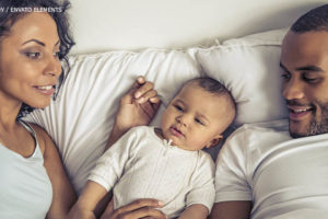 Imagem Acessível: casal composto por uma mulher e um homem, ambos de peles negras, estão deitados em uma cama cuidando de um bebê que está entre eles.